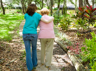 We see the backs of two people walking side-by-side - a young student and an older woman. The student supports the elder woman as they walk along a wooded path.