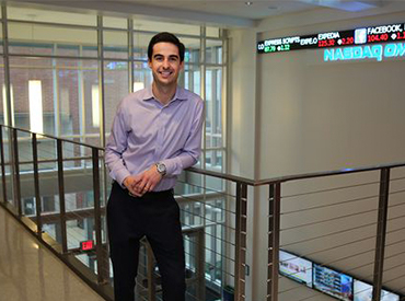 A graduate student in a button-down shirt and dress slacks stands in front of the stock ticker in the business center